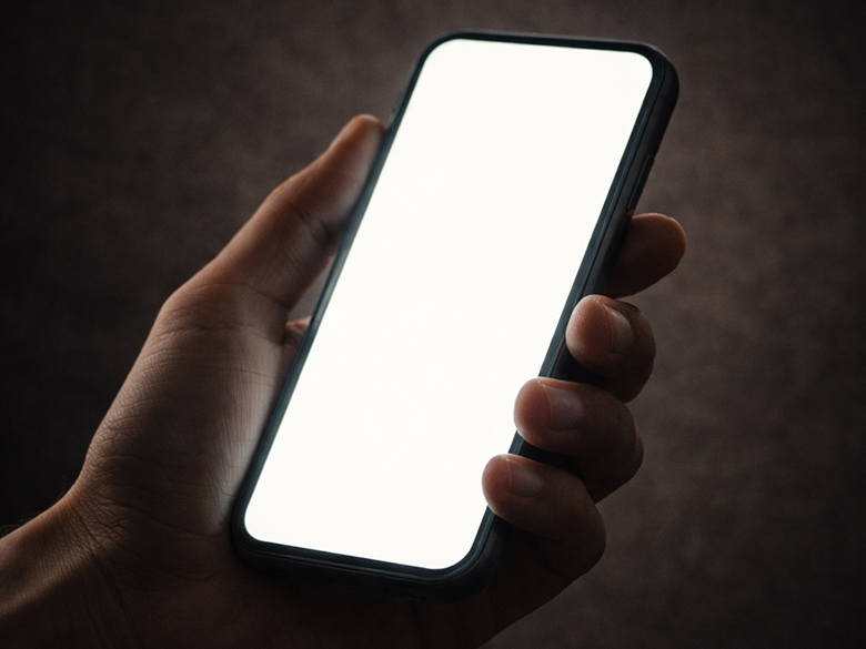 Close-up of a hand holding a smartphone in a dark, textured space, the screen casting a soft glow while the surrounding environment fades into shadow.